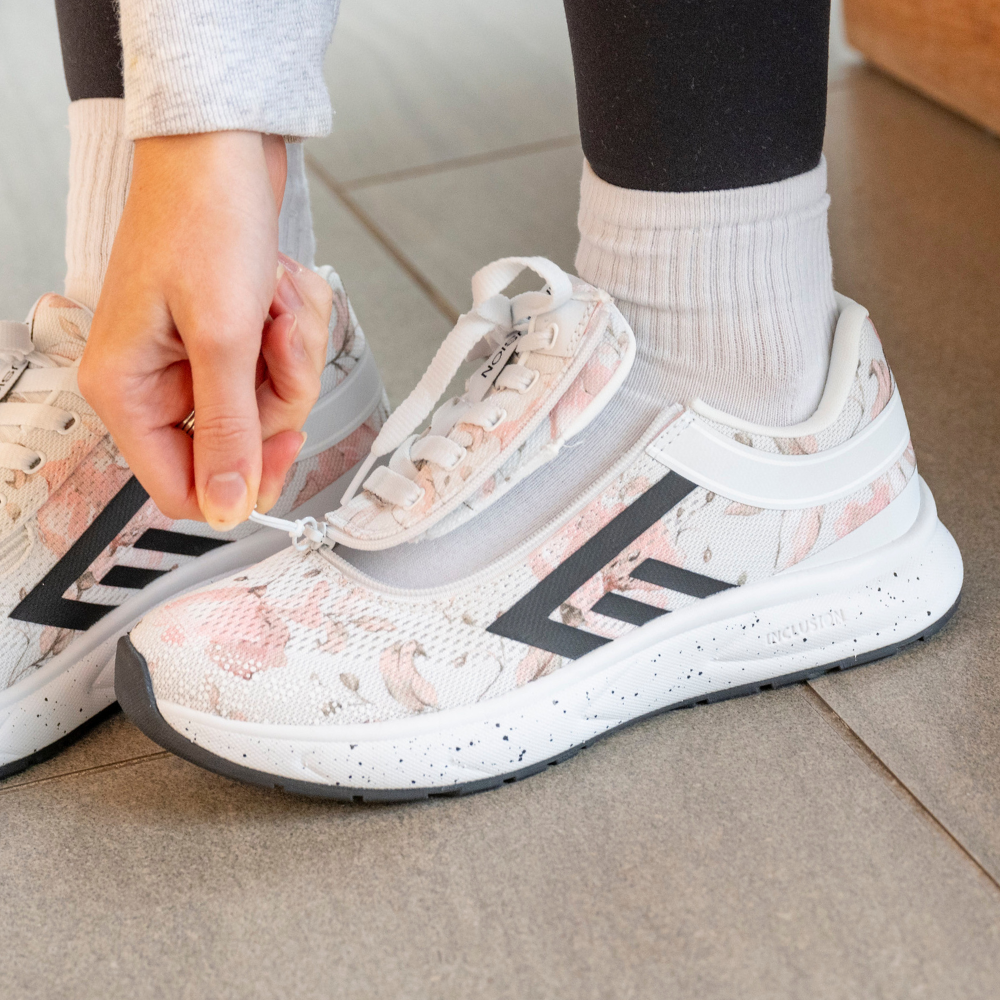 A person wearing white sneakers with pink floral patterns and black stripes is pulling up a zipper on the shoe, standing on a tiled floor.