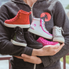 A person in a grey hoodie holds five pairs of small, colorful sneakers—red, black, white with blue and pink accents, pink, and gray—cradled in their arms.