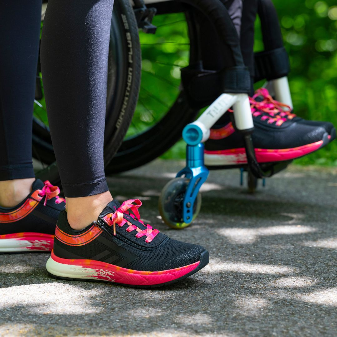 Two people wearing black athletic shoes with pink laces are outdoors; one person is standing, and the other is seated in a wheelchair. Green foliage is visible in the background.