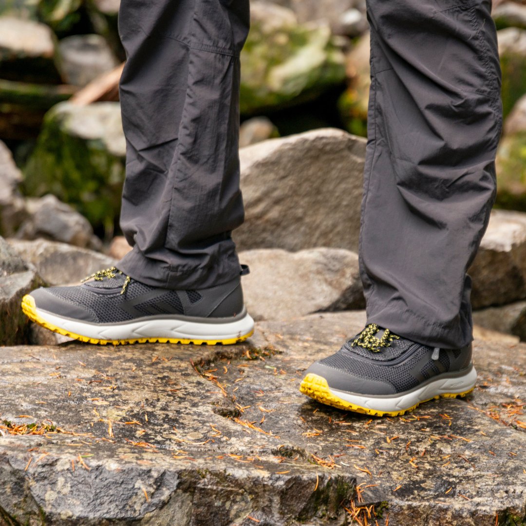Male standing on wet rock wearing rain pants and sport inclusion trail shoes. Additional boulders in background.