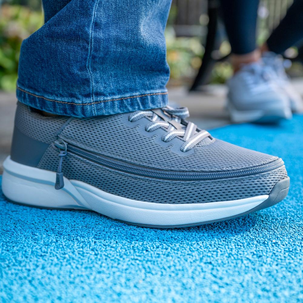 Close-up of a person wearing grey athletic shoes and blue jeans, standing on a textured blue surface. Another person in similar shoes is blurred in the background.