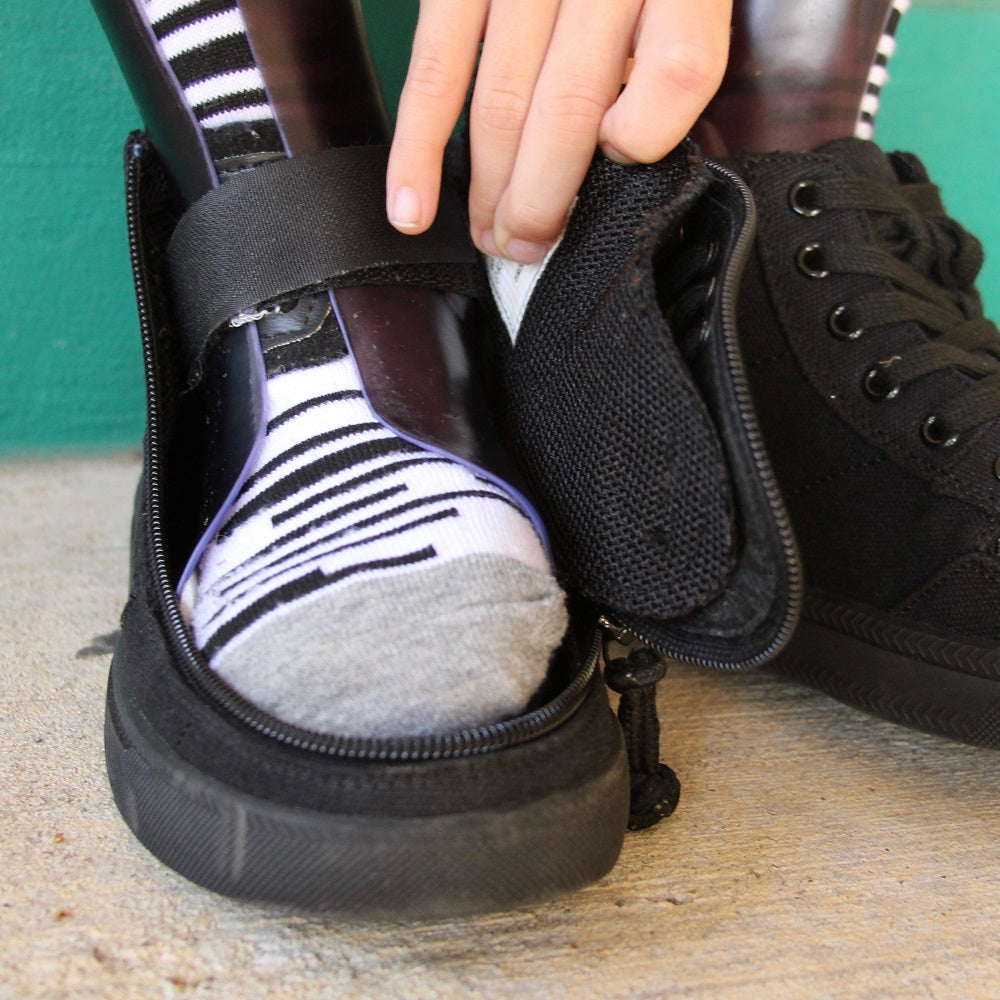 A hand helps put on a black shoe with a wide opening and zipper, revealing a striped sock and foot with orthotic brace inside, on a concrete surface with a teal wall in the background.