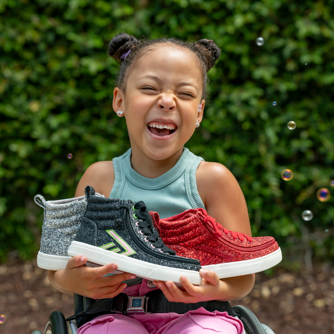 A smiling child in a wheelchair holds up two glittery high‑top sneakers, surrounded by bubbles and greenery.