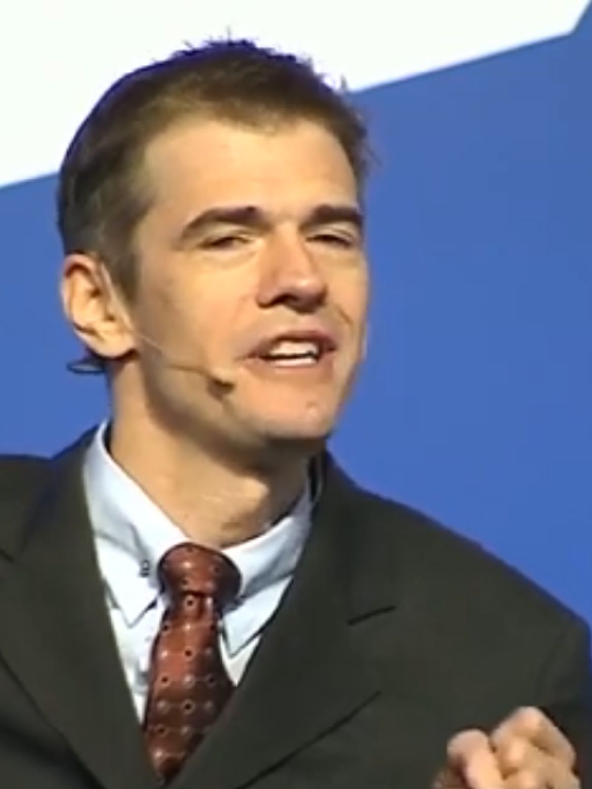 Man in a suit and tie speaking in front of a blue background