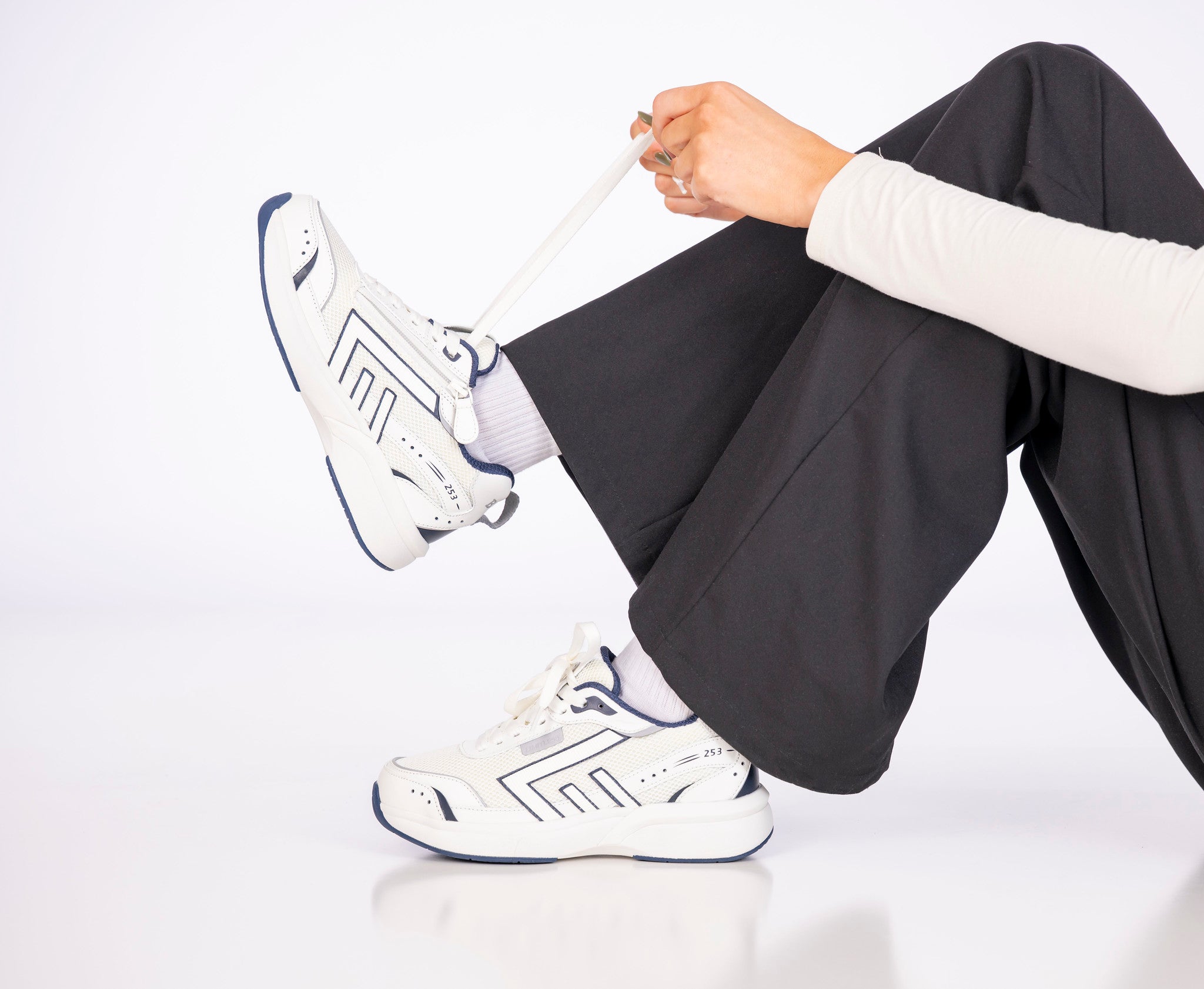 Person wearing white sneakers with a brand logo, sitting on a white surface.