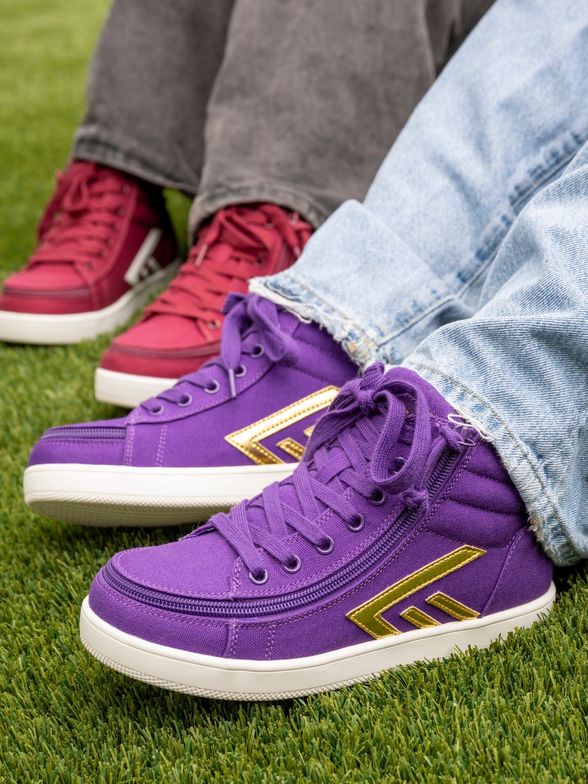 Three pairs of casual sneakers shown on feet sitting on green grass. The foreground pair is purple with a gold geometric side logo and white sole, worn with light blue jeans.