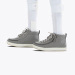 A close-up side view of a person wearing dark grey BILLY Classic D|R II High shoes from BILLY Footwear and white ankle-foot orthoses over white socks, standing on a light background.