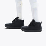 A person with white prosthetic legs wears Black to the Floor BILLY D|R II Short Wrap High sneakers by BILLY Footwear, featuring removable insoles, and white socks, standing on a light background.
