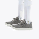 A side close-up of a person wearing the SALE - Dark Grey BILLY D|R II Short Wrap Low orthotic shoes by BILLY Footwear with white leg braces, set against a plain white background.