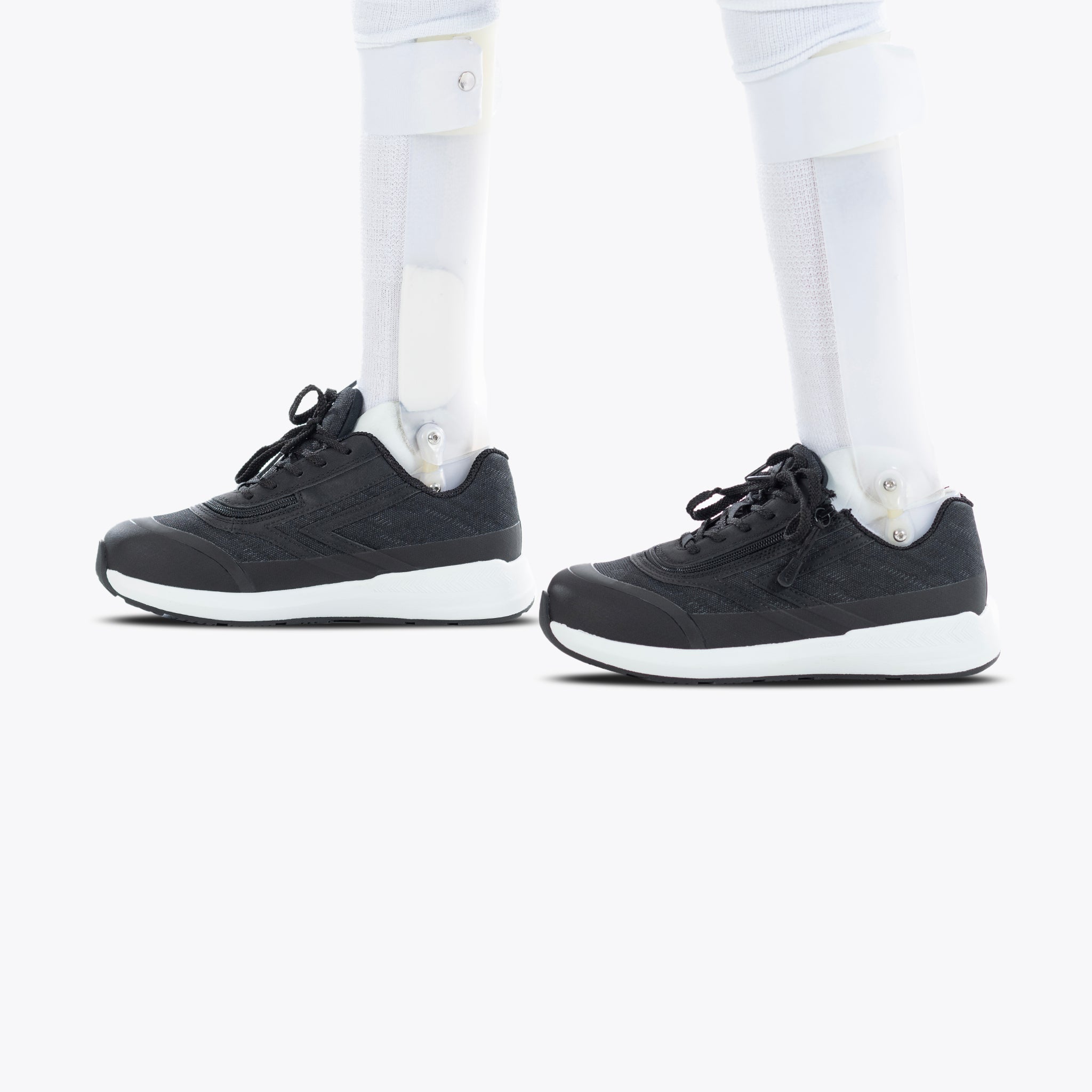 A person with prosthetic legs wears Black BILLY Goat orthotic shoes by BILLY Footwear, featuring white soles, standing on a plain white background.