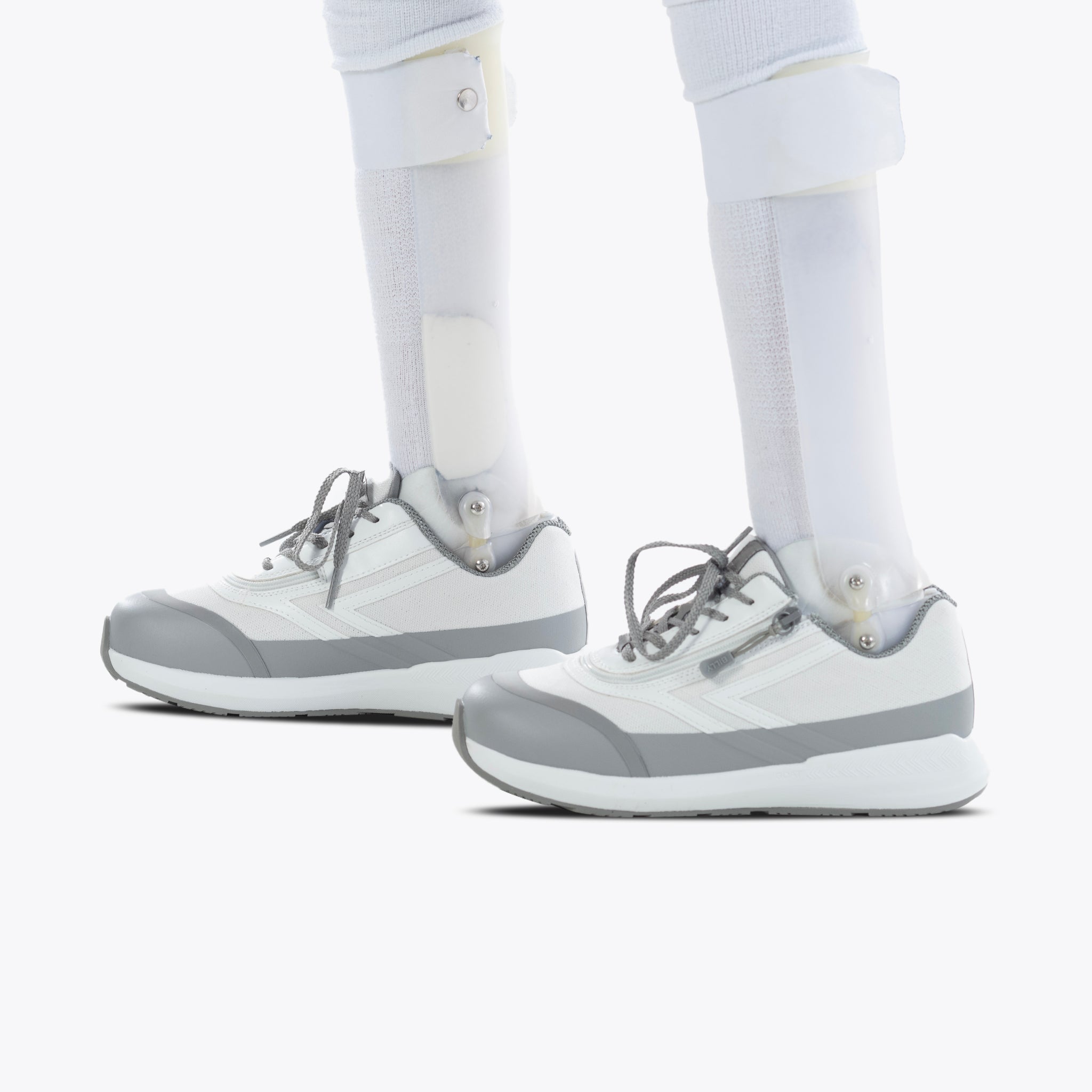 Close-up of two prosthetic legs fitted with SALE - White BILLY Goat shoes by BILLY Footwear, featuring an easy on-and-off zipper, standing on a white background.