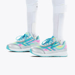A person wearing white leg braces and Rainbow Tie Dye BILLY Goat sneakers by BILLY Footwear—adaptive, AFO-friendly kids’ shoes with turquoise laces, silver accents, and blue soles—stands against a white background.