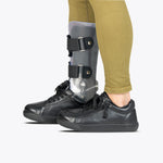 A person wearing tan pants and black BILLY Footwear SALE - Black to the Floor Leather BILLY Classic D|R II Low shoes, designed for AFOs, with an orthopedic leg brace on their right lower leg, stands against a white background.
