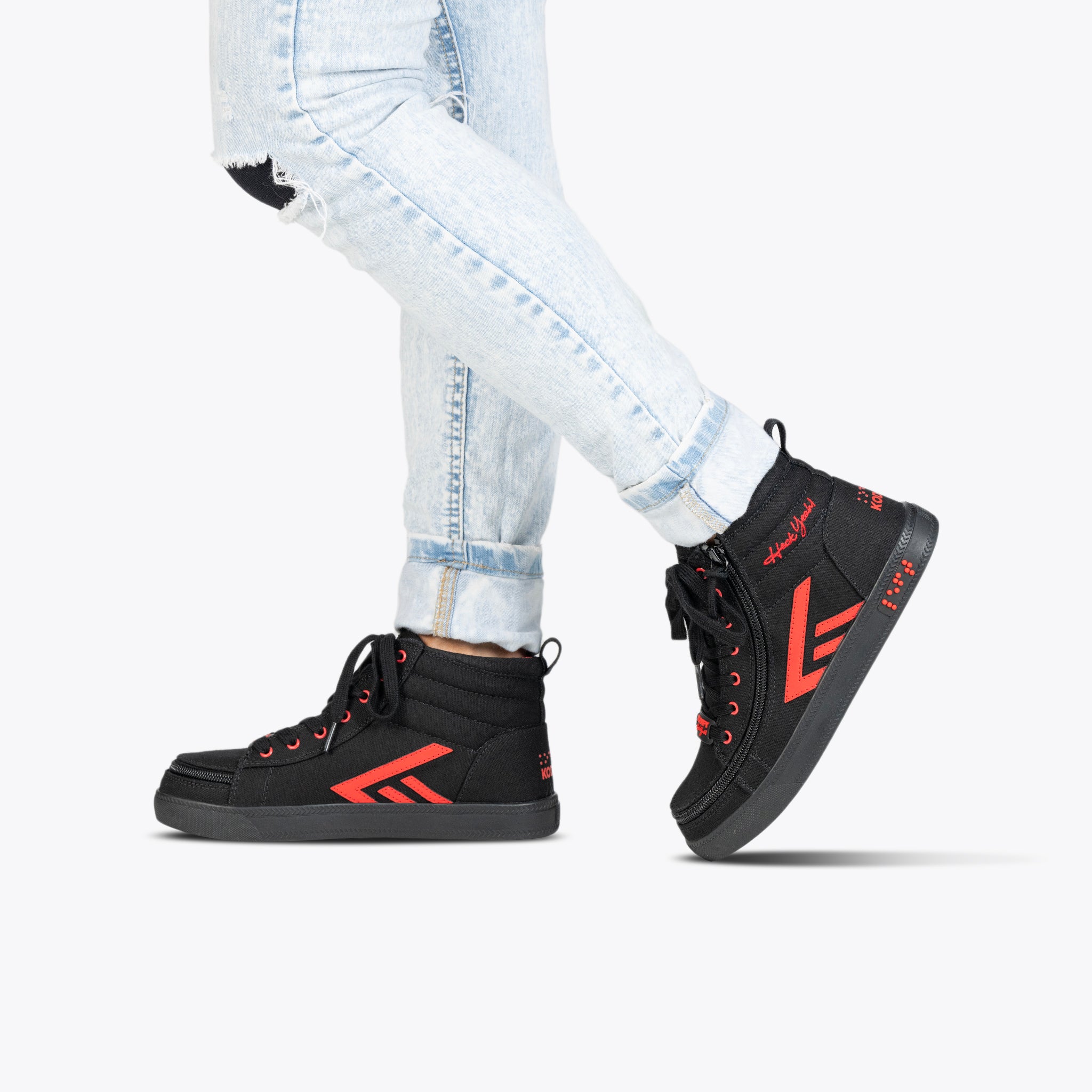 A person stands on tiptoe in light blue ripped jeans and SALE - Kodify Black BILLY CS High sneakers by BILLY Footwear, featuring red accents and laces, against a plain white background.