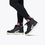 A person wears BILLY Footwear's Black Glitz BILLY CS High with white soles, black leggings, and black socks with a pink floral design, standing against a plain white background—stylish kids’ skate shoes with sparkle.