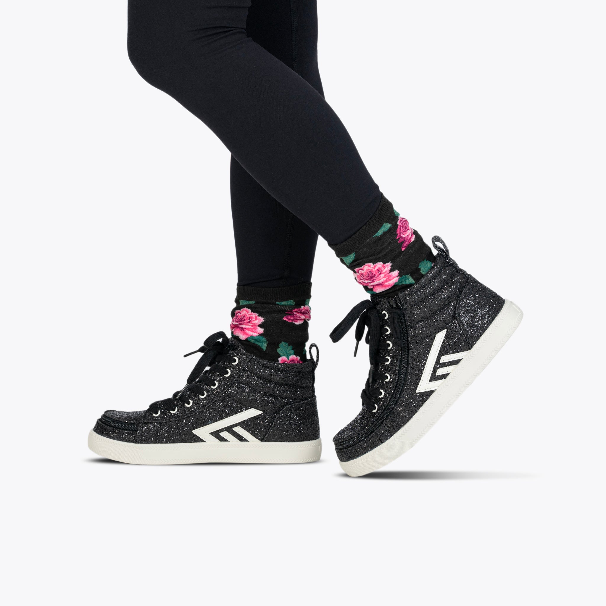 A person wears BILLY Footwear's Black Glitz BILLY CS High with white soles, black leggings, and black socks with a pink floral design, standing against a plain white background—stylish kids’ skate shoes with sparkle.