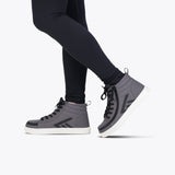 A person in black leggings and Charcoal/Black BILLY CS High sneakers by BILLY Footwear, featuring a full wrap zipper, stands on one leg with the other raised and bent at the knee against a plain white background.