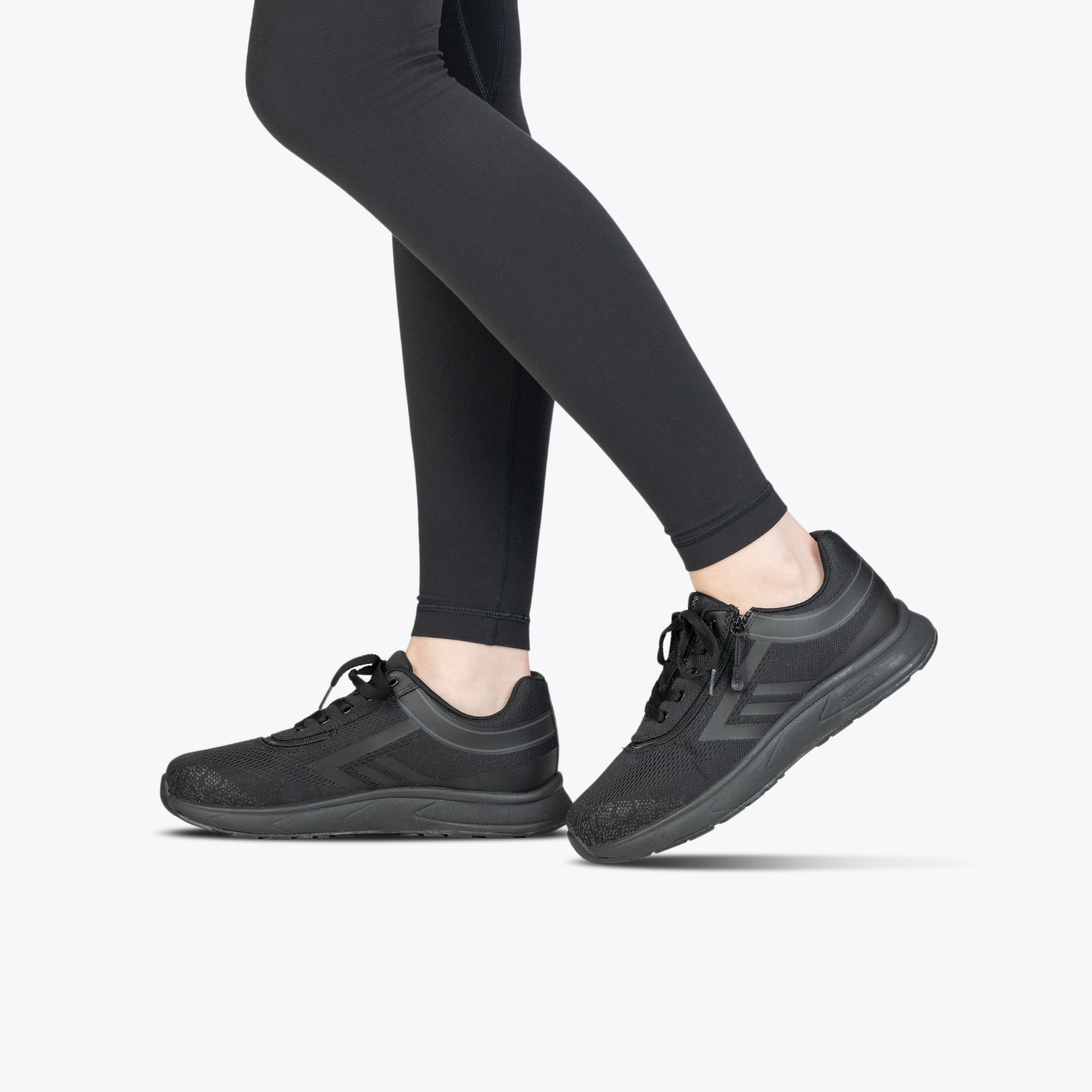 A person wearing Black to the Floor BILLY Sport Inclusion shoes from BILLY Footwear and black leggings stands on a light gray background, with only their legs and feet visible.