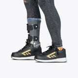 Side view of a person wearing black jeans, a metal leg brace on their left leg, and BILLY Footwear’s Black/Gold BILLY Goat Hoop shoes, standing on a light gray background.