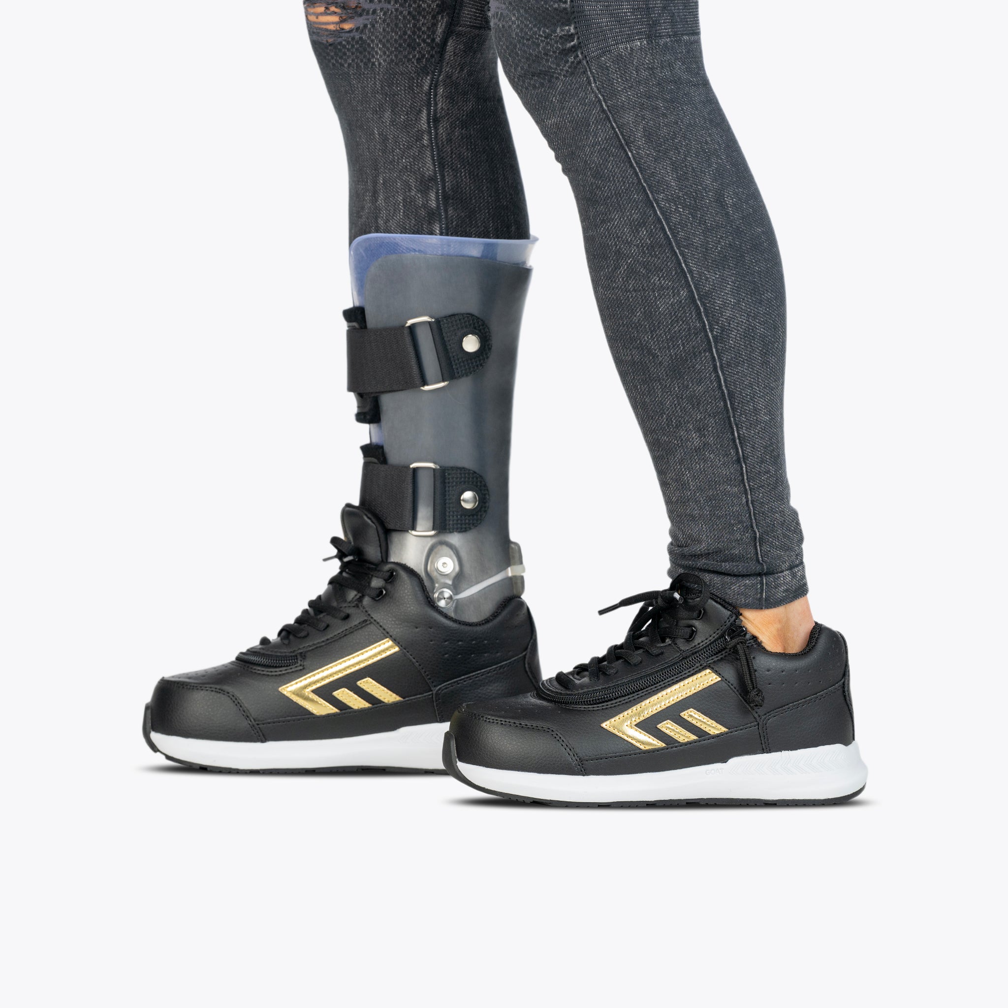 Side view of a person wearing black jeans, a metal leg brace on their left leg, and BILLY Footwear’s Black/Gold BILLY Goat Hoop shoes, standing on a light gray background.