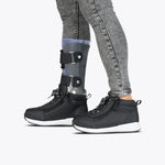 A child wearing gray jeans and black Kids'/Toddler BILLY Goat DZ High sneakers by BILLY Footwear with a visible left prosthetic leg. The shoes are orthotic-friendly, set against a plain, light background.