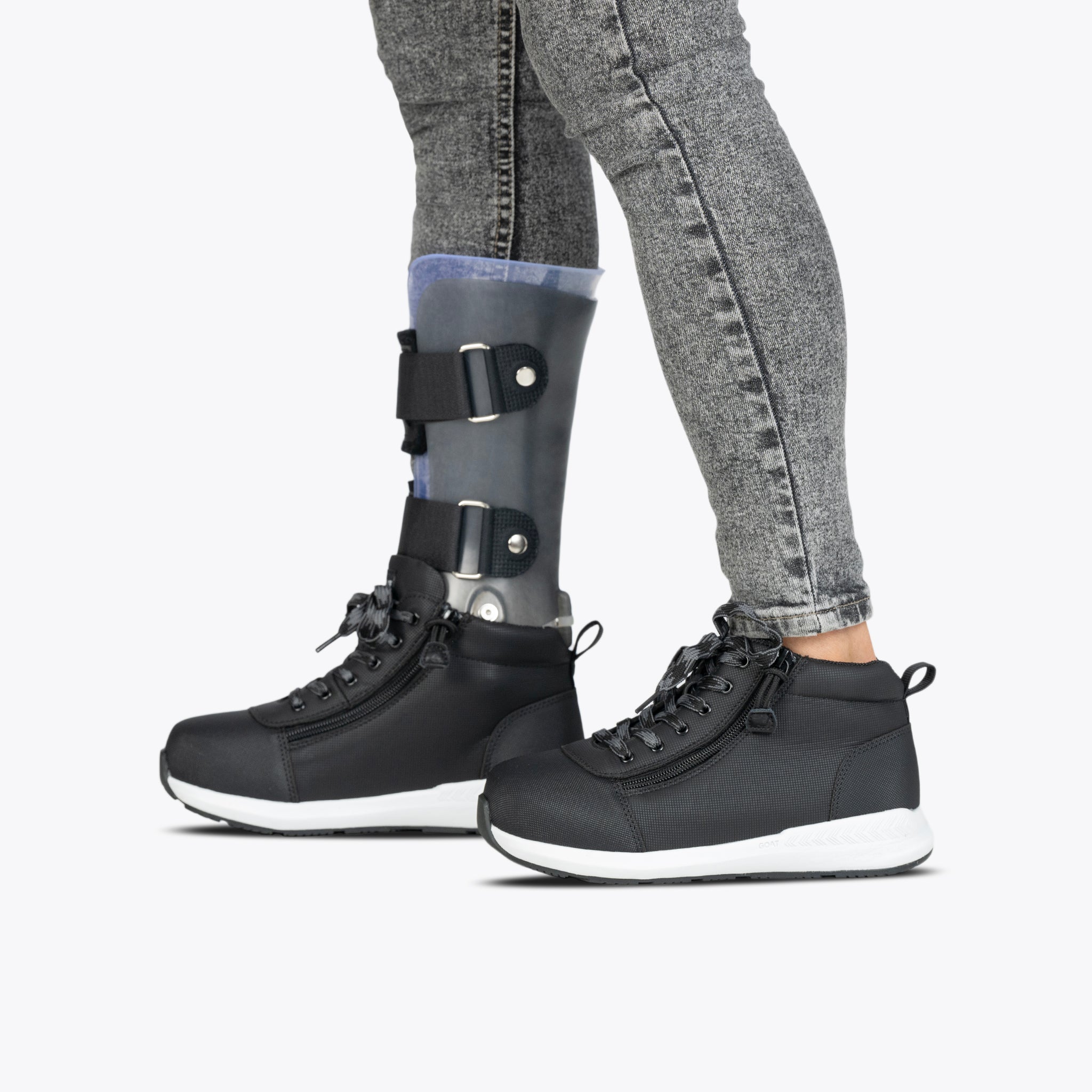 A child wearing gray jeans and black Kids'/Toddler BILLY Goat DZ High sneakers by BILLY Footwear with a visible left prosthetic leg. The shoes are orthotic-friendly, set against a plain, light background.