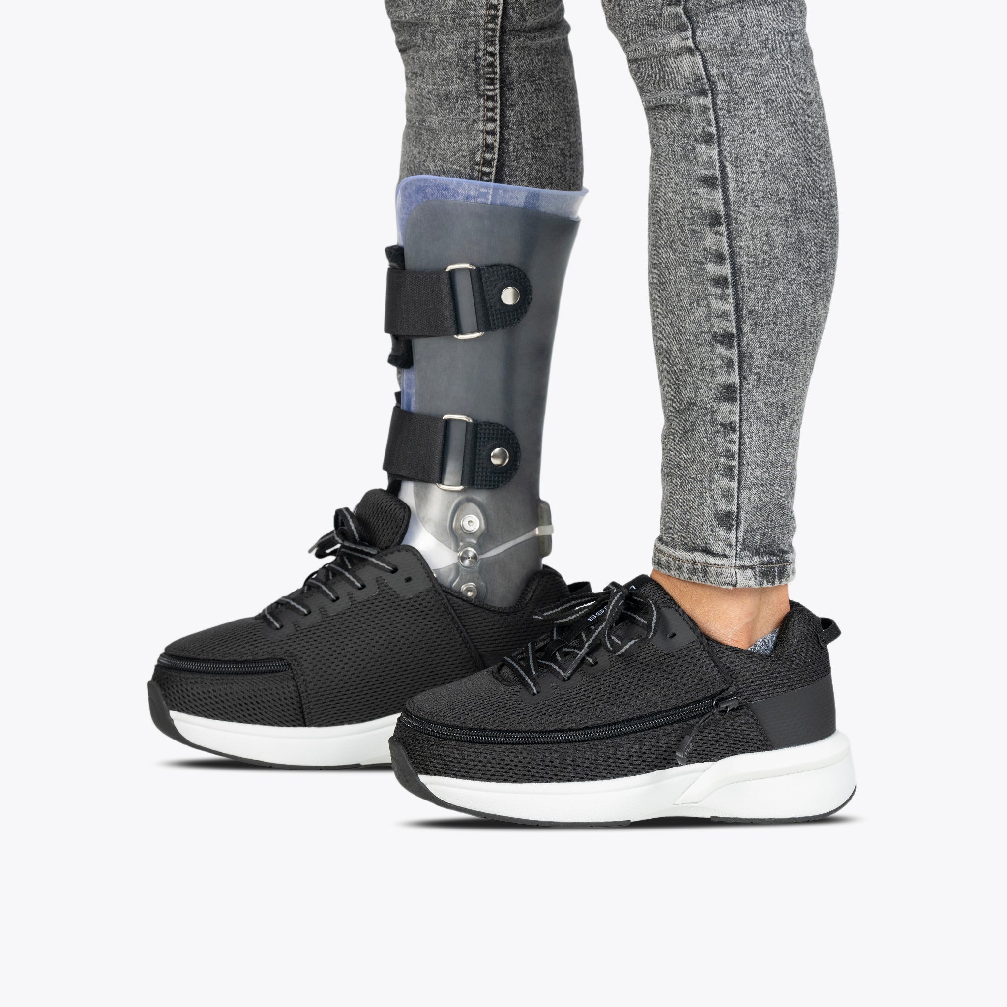 A person in grey jeans, Black BILLY Limitless shoes by BILLY Footwear, and a black orthopedic leg brace on their left leg stands against a plain white background.