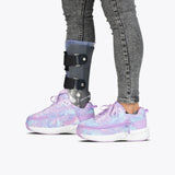 A person wearing gray jeans and mismatched BILLY Footwear Lilac Watercolor BILLY Limitless adaptive shoes, with a visible prosthetic leg brace on the left leg, stands against a plain white background.