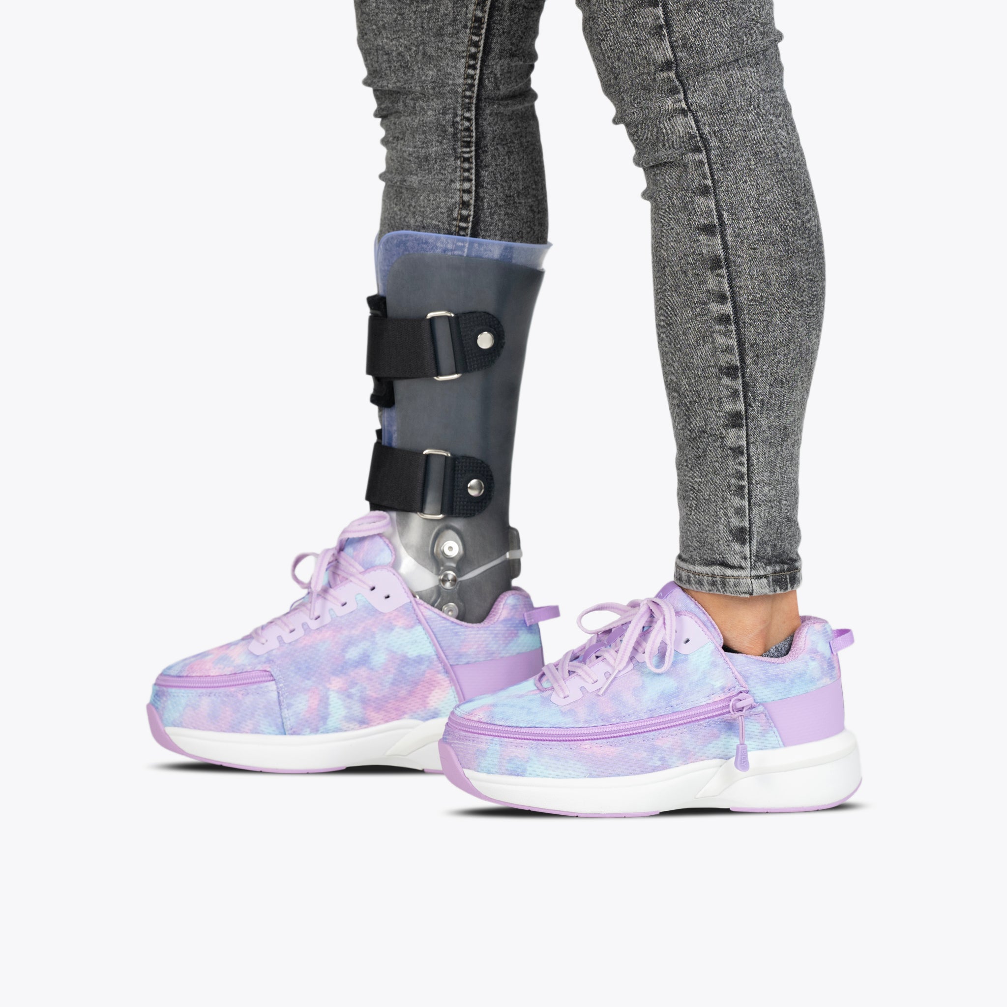 A person wearing gray jeans and mismatched BILLY Footwear Lilac Watercolor BILLY Limitless adaptive shoes, with a visible prosthetic leg brace on the left leg, stands against a plain white background.