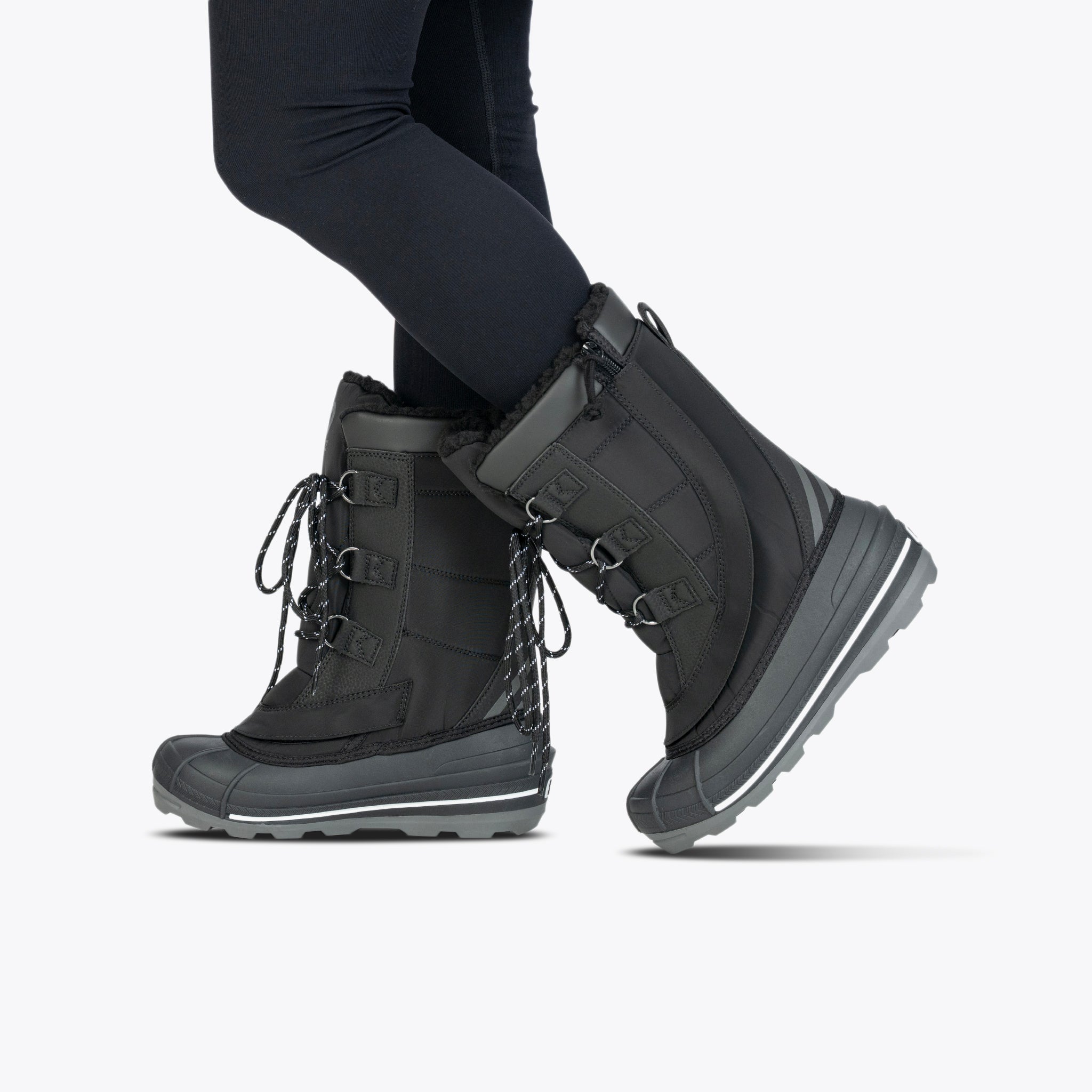 A person wearing Black BILLY Ice Boot by BILLY Footwear, featuring thick soles, fur lining, and lace-up fronts, is paired with black leggings and stands against a plain light background.