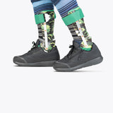 Two lower legs wearing BILLY Footwear Men's Black to the Floor BILLY Goat shoes and camouflage-patterned orthotic leg braces with green straps, over striped socks and blue clothing, set against a plain white background.