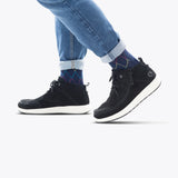 A person wearing rolled-up blue jeans, colorful argyle socks, and the Men's Black Suede BILLY Comfort Chukka by BILLY Footwear with white soles walks on a light gray background.