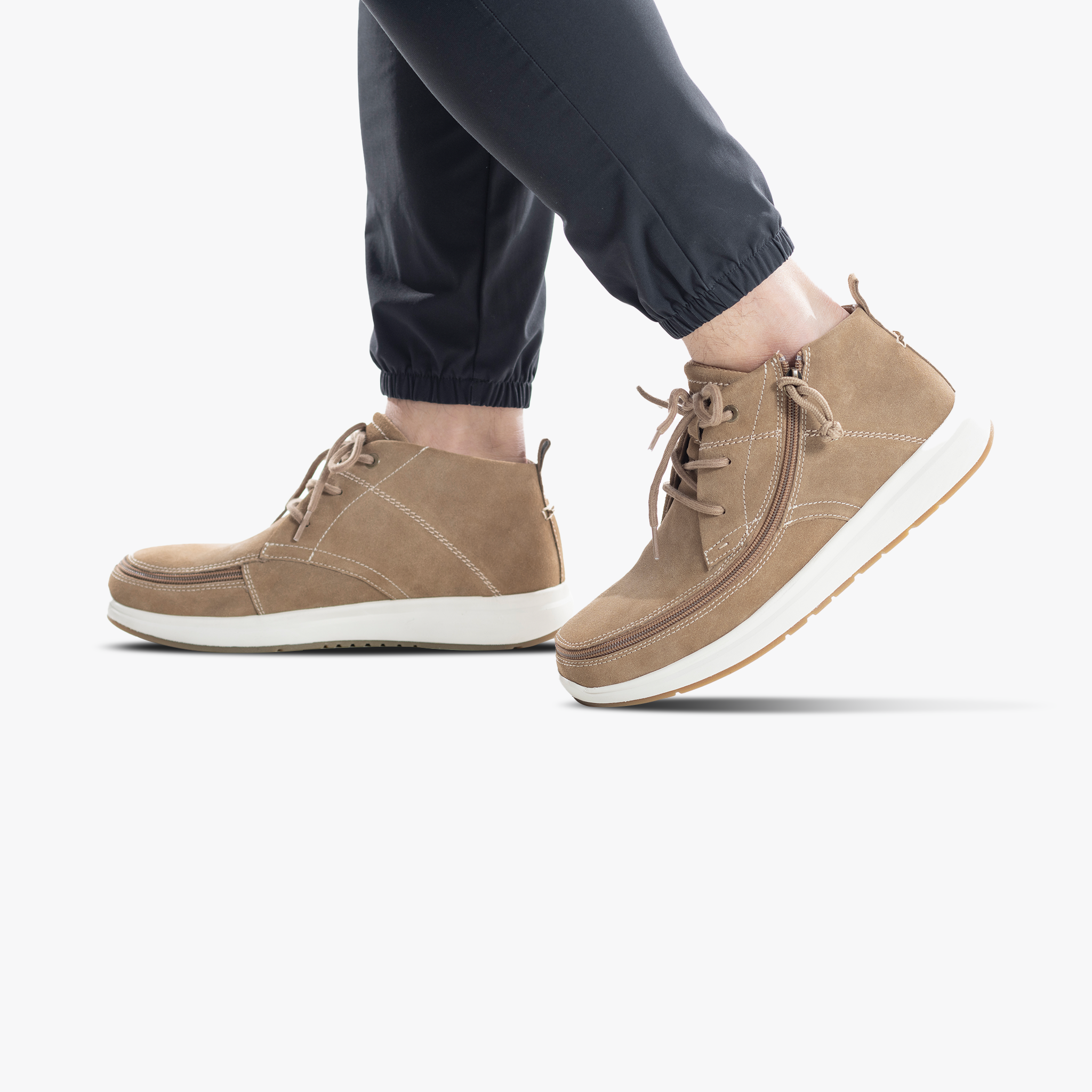 A person wearing Men's Sand Suede BILLY Comfort Chukka shoes by BILLY Footwear with white soles and dark jogger pants stands on a white background.