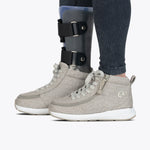 Wearing BILLY Footwear's Women's Grey Jersey BILLY Goat Classic High shoes and an orthotic leg brace over striped leggings, a person stands side by side from the knee down on a white background.