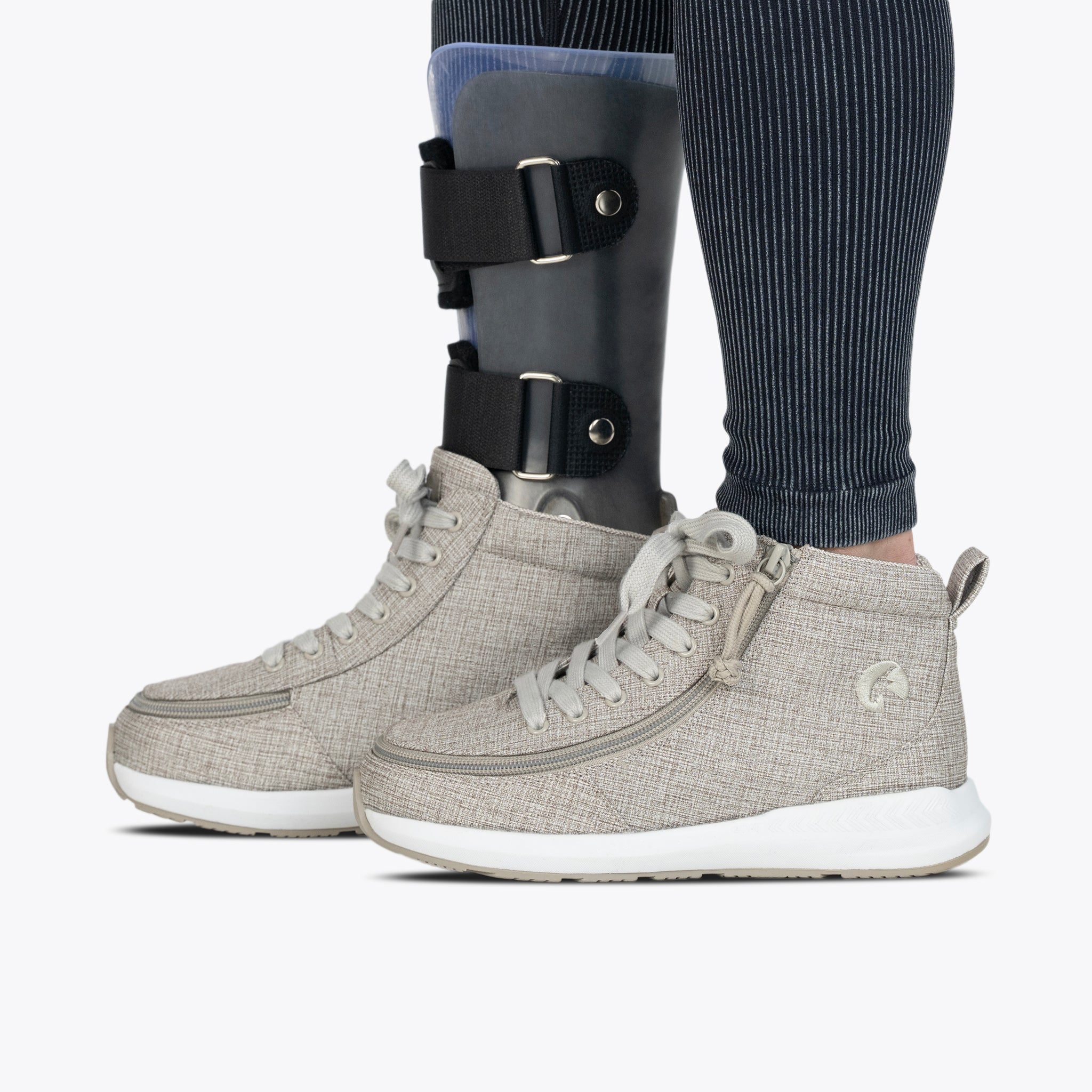 Wearing BILLY Footwear's Women's Grey Jersey BILLY Goat Classic High shoes and an orthotic leg brace over striped leggings, a person stands side by side from the knee down on a white background.