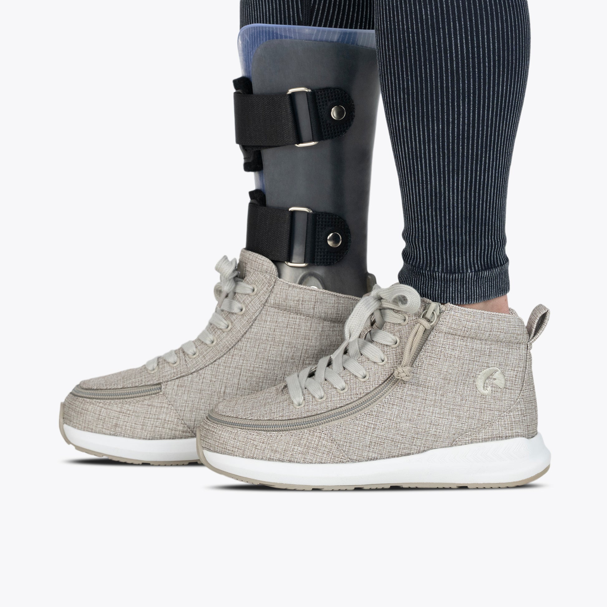Wearing BILLY Footwear's Women's Grey Jersey BILLY Goat Classic High shoes and an orthotic leg brace over striped leggings, a person stands side by side from the knee down on a white background.