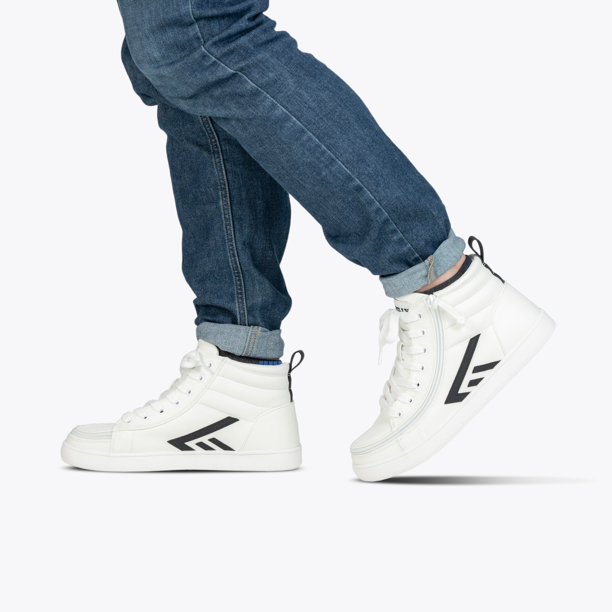 A person is shown walking against a plain white background, wearing rolled-up blue jeans and the Men's White/Black BILLY CS High sneakers from BILLY Footwear, which feature black geometric designs and a signature full wrap zipper.