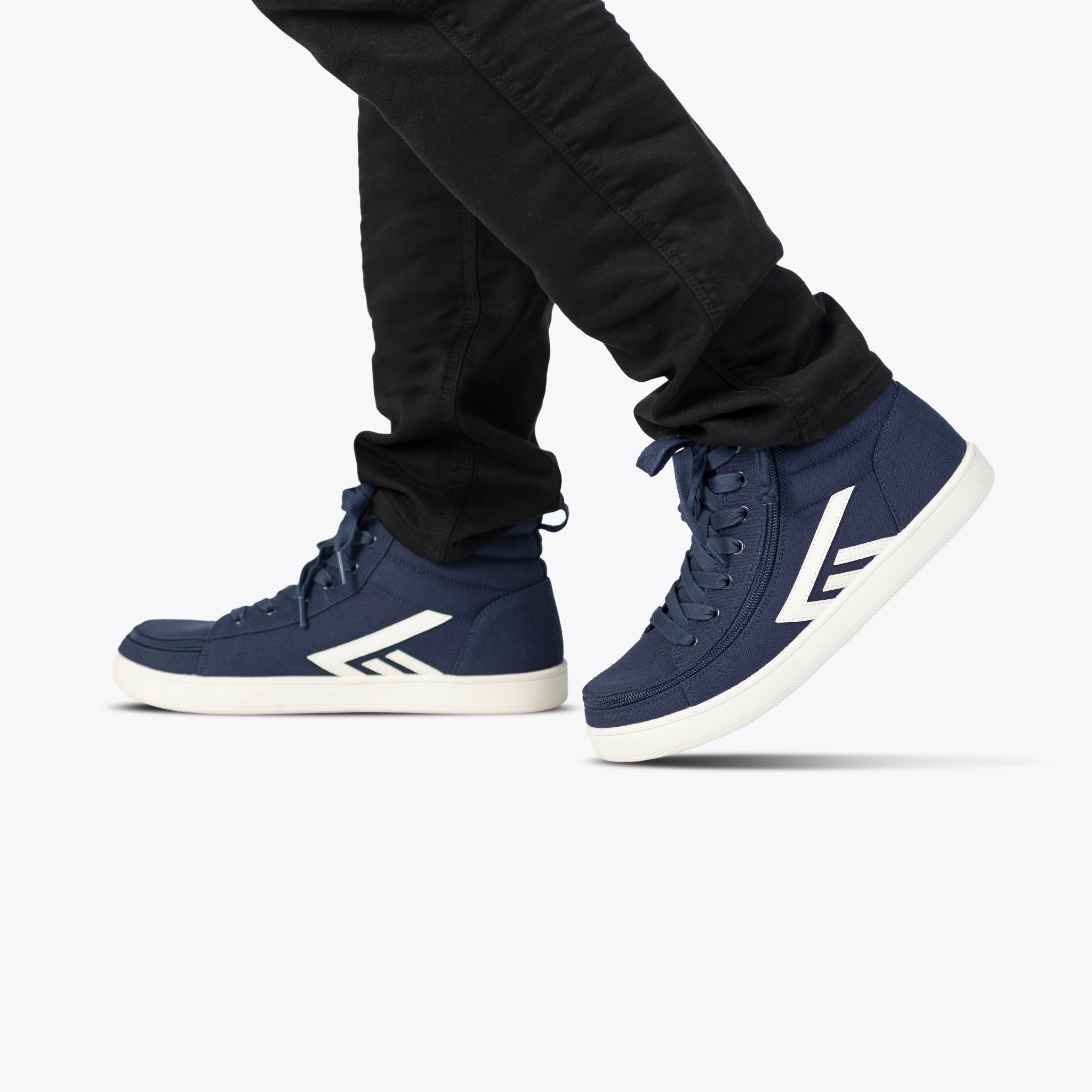 A person in black pants walks against a plain white background wearing BILLY Footwear's Men's Navy/White BILLY CS High sneakers, featuring a white sole and the signature L logo.