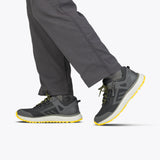 A person wears Men's Charcoal/Yellow BILLY Inclusion Trail sneakers from BILLY Footwear, featuring yellow soles and accents, paired with dark gray pants. Only their lower legs and feet are visible against a light background.