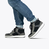 A person wearing BILLY Footwear's Men's Black/Charcoal/Black BILLY Comfort DZ High sneakers and blue jeans walks with one foot slightly raised against a plain white background, highlighting these comfortable shoes.