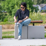 A man in sunglasses and casual clothes sits outdoors on a concrete bench, tying his BILLY Footwear Men's White/Grey/Black Comfort DZ High sneakers. Green trees, grass, and a blurred view of houses complete the relaxed scene.