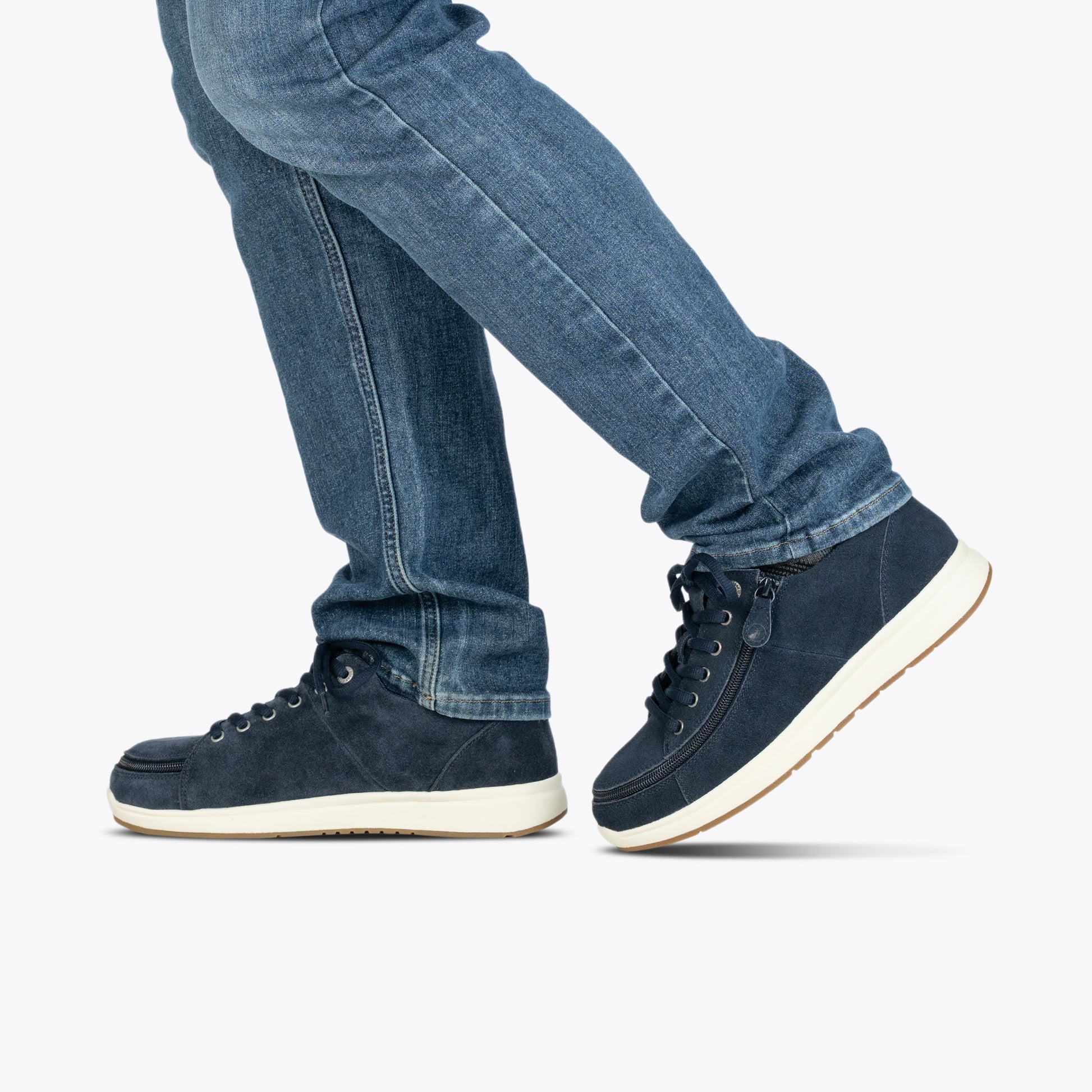 A person in blue jeans and BILLY Footwear Men's Navy Suede BILLY Comfort Plush shoes with white soles walks, shown from the knees down against a plain white background.