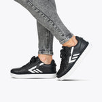 A person wears the Women's Black/White BILLY CS Low by BILLY Footwear—black uppers, white soles, stripe accents—paired with gray skinny jeans against a plain light background. These sensory-friendly sneakers combine style and comfort.