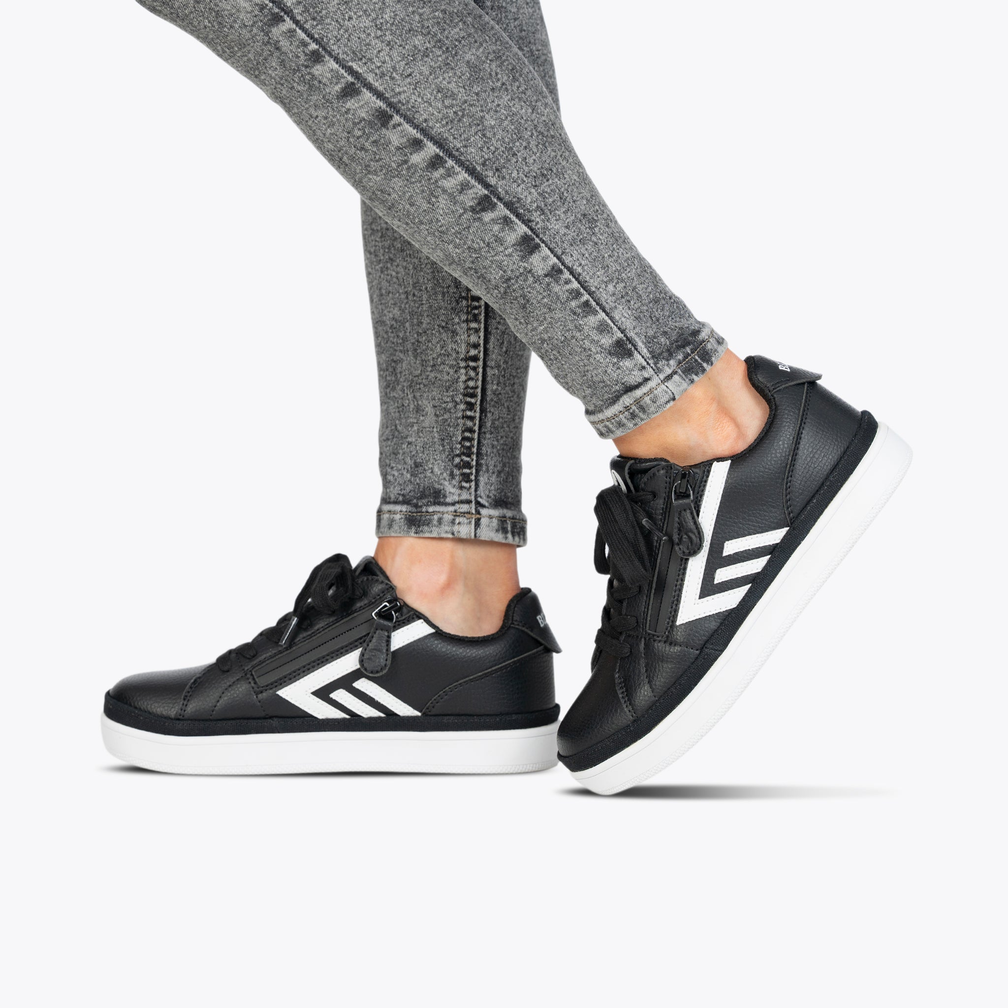 A person wears the Women's Black/White BILLY CS Low by BILLY Footwear—black uppers, white soles, stripe accents—paired with gray skinny jeans against a plain light background. These sensory-friendly sneakers combine style and comfort.