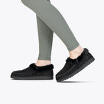 A person stands on tiptoes against a plain white background, showing only their lower legs in light olive green leggings and wearing BILLY Footwear Women's Black BILLY Cozy Slippers with a full wrap zipper.