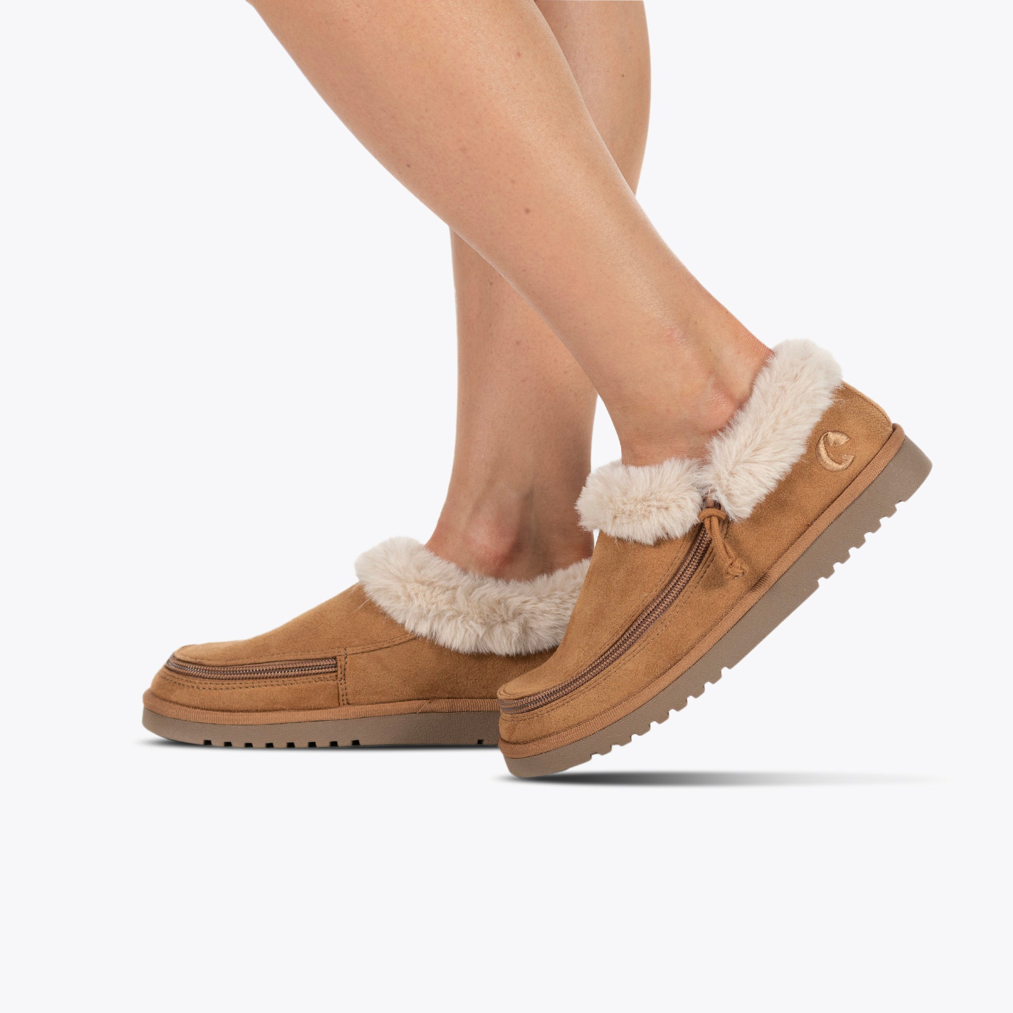 A person wears the Women's Chestnut BILLY Cozy Slipper by BILLY Footwear, featuring plush cream faux fur lining and side wrap zippers, while standing on tiptoe against a plain white background.