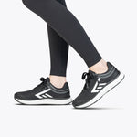 A person stands on tiptoe against a plain white background, wearing black athletic leggings and Women's Black/White BILLY Sport Inclusion sneakers by BILLY Footwear, featuring white soles and geometric designs.