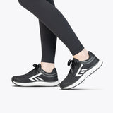 A person stands on tiptoe against a plain white background, wearing black athletic leggings and Women's Black/White BILLY Sport Inclusion sneakers by BILLY Footwear, featuring white soles and geometric designs.