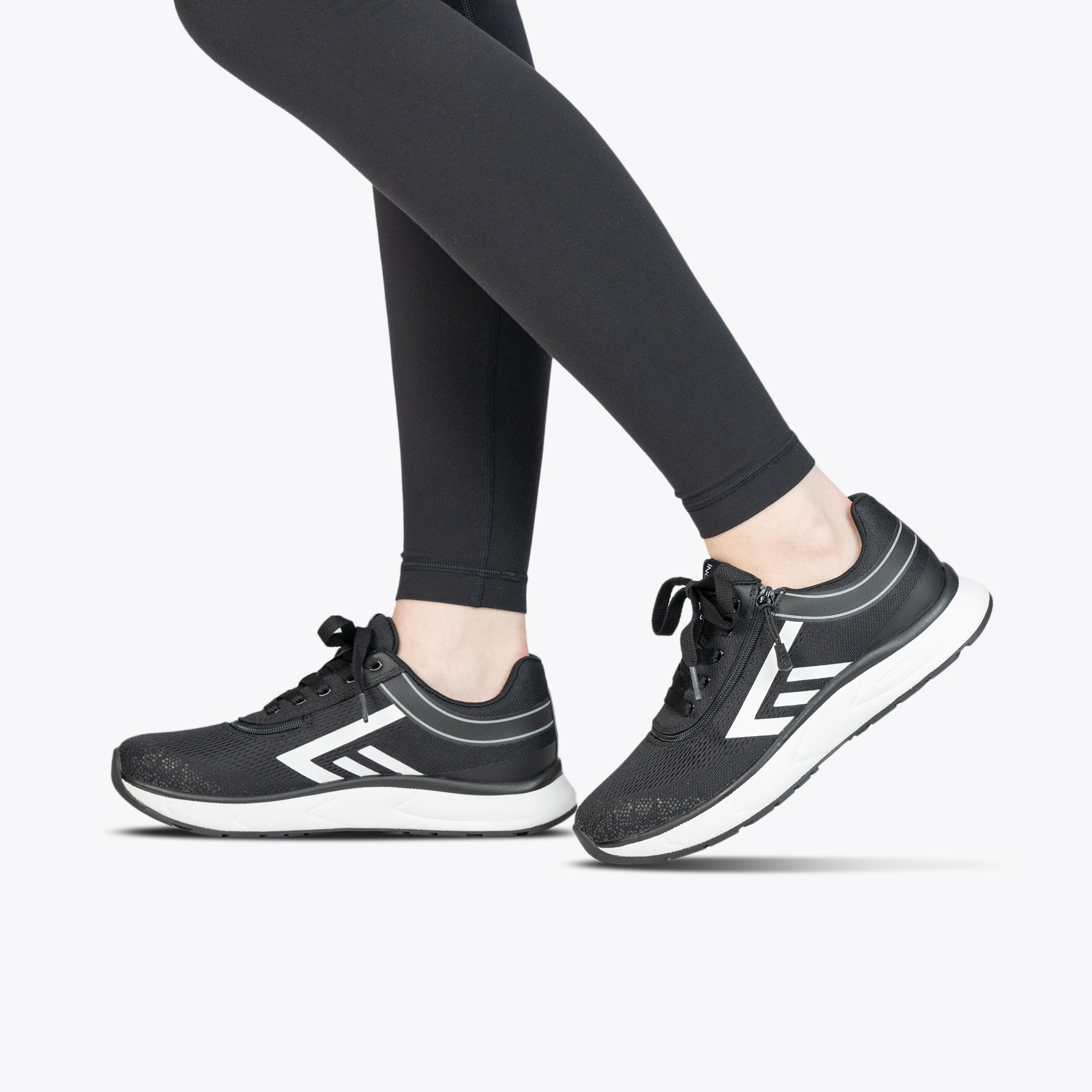 A person stands on tiptoe against a plain white background, wearing black athletic leggings and Women's Black/White BILLY Sport Inclusion sneakers by BILLY Footwear, featuring white soles and geometric designs.