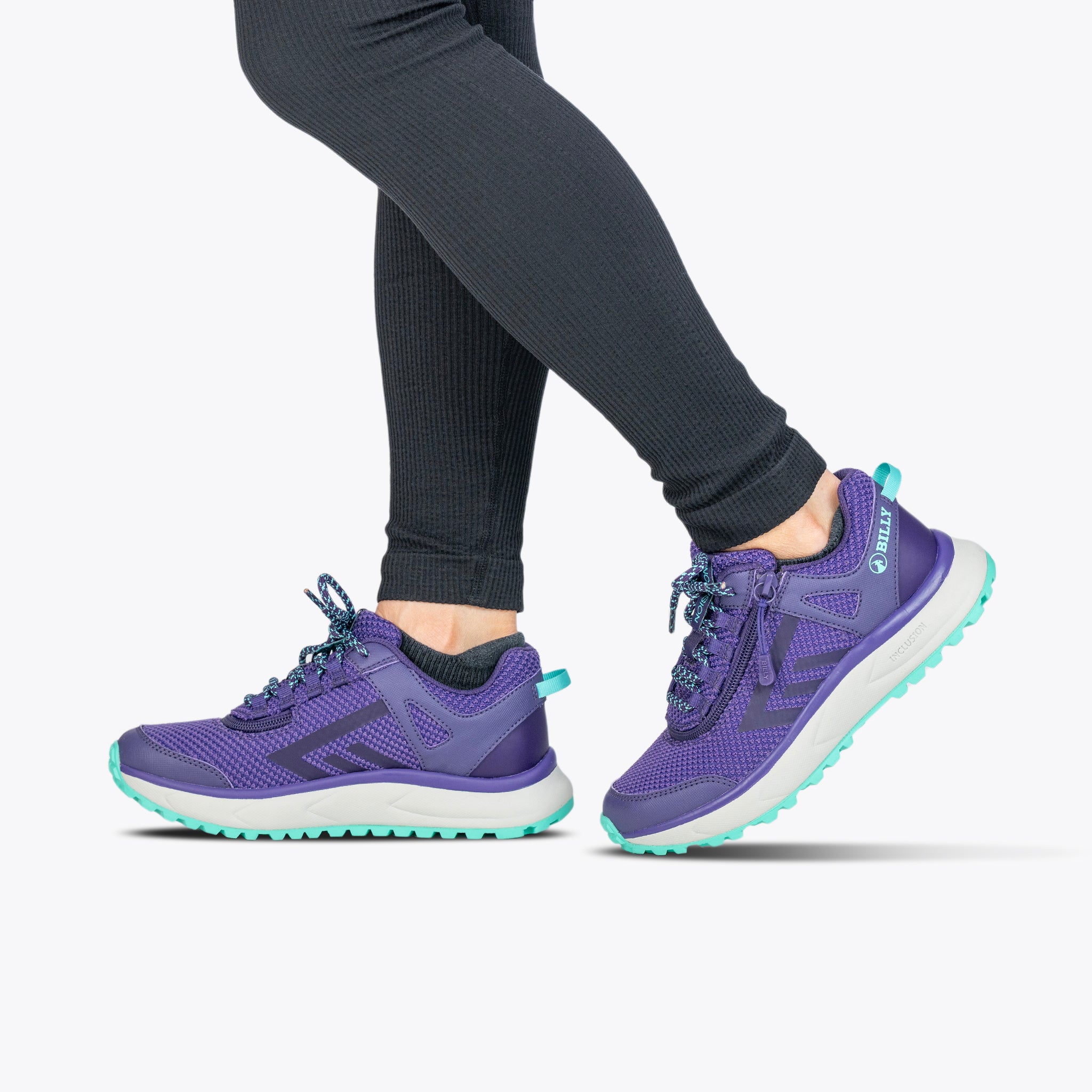 A person in black leggings and Women's Purple/Mint BILLY Inclusion Trail sneakers by BILLY Footwear, featuring an easy on-off zipper, walks against a plain white background.